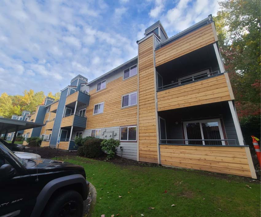 Low-angle exterior view of a multi-story apartment building featuring a mix of natural wood lap siding and blue-gray accent panels on the balconies.